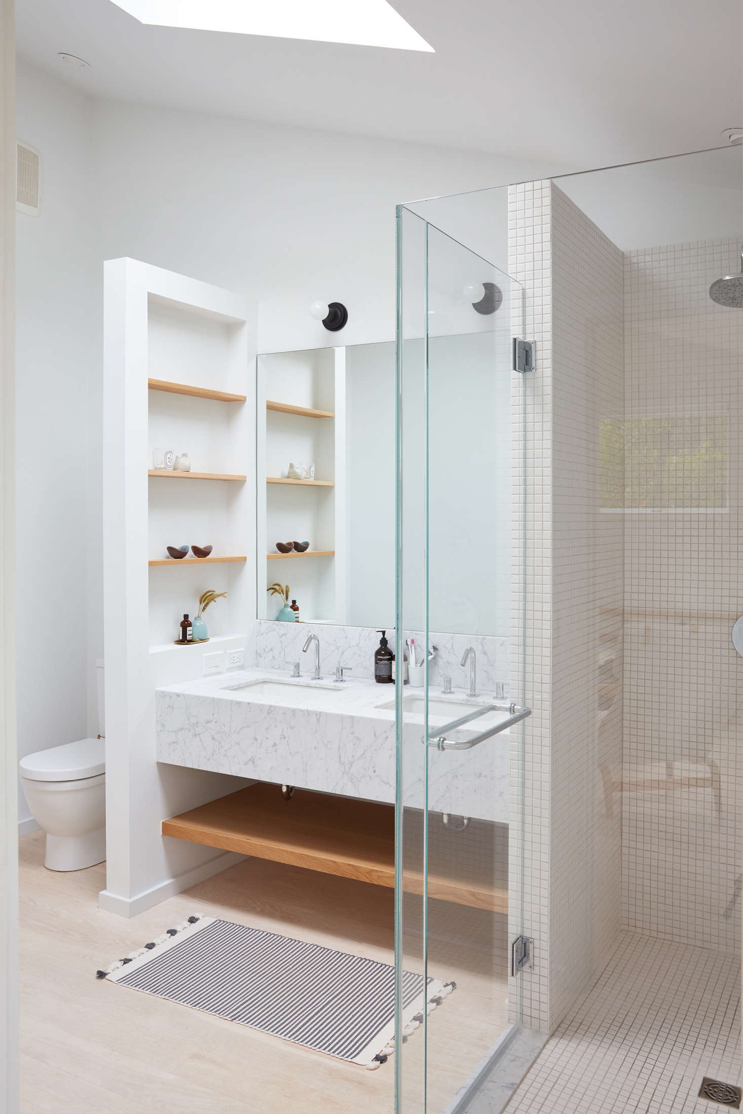 In the master bedroom, a custom marble vanity (also with marble from ABC Stone in Brooklyn) is fitted with custom oak shelves, perfect for storing bath essentials. A glass-enclosed shower uses square tiles from Nemo Tile. The toilet is the Duravit Happy D., and the striped rug is from Nickey Keyhoe in LA.