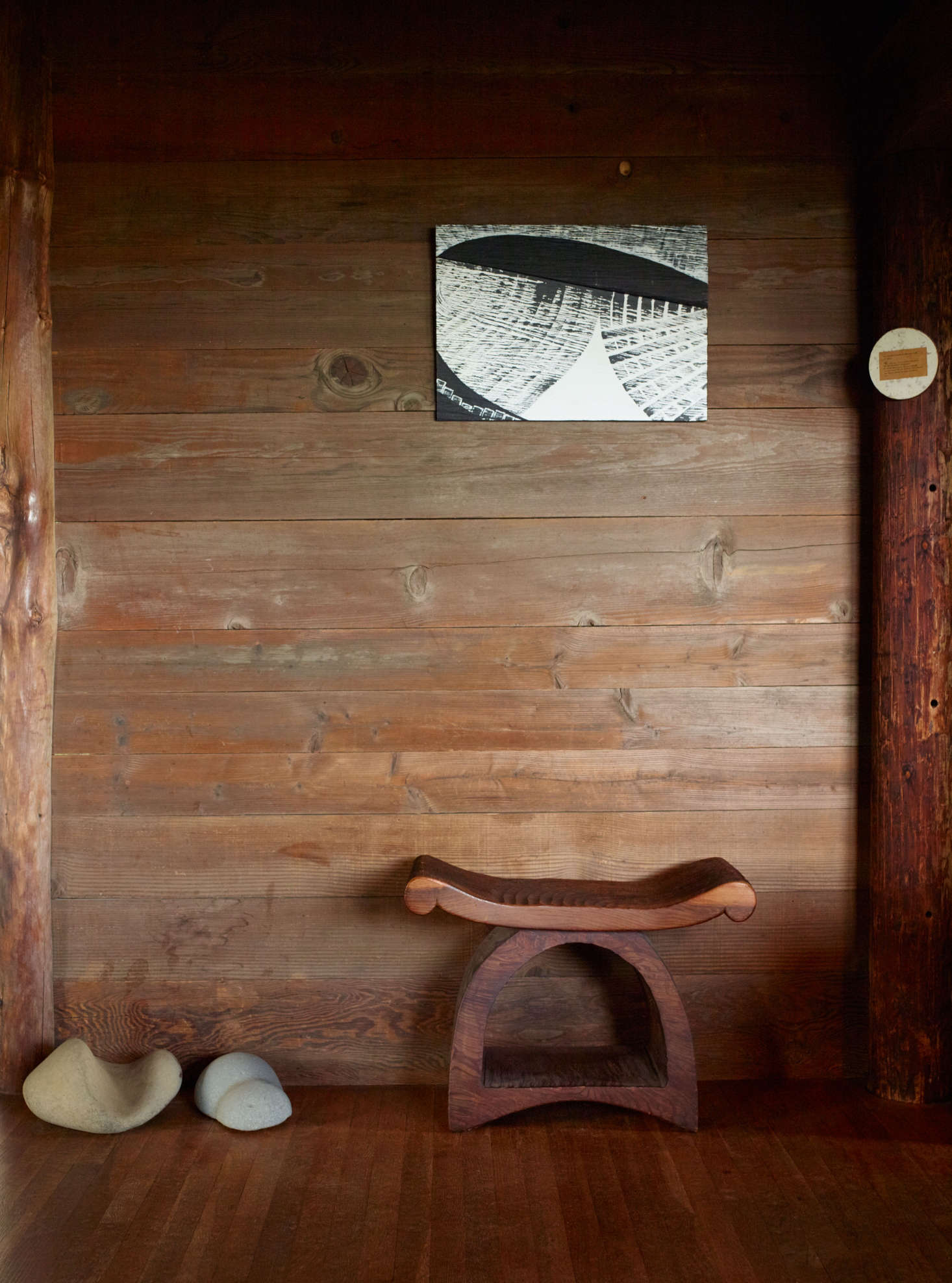 Blunk’s painting in black and white mirrors the chainsaw markings on the underlying redwood plank. The flared and curved outlines of the redwood stool echo the silhouettes on a display of river rocks.