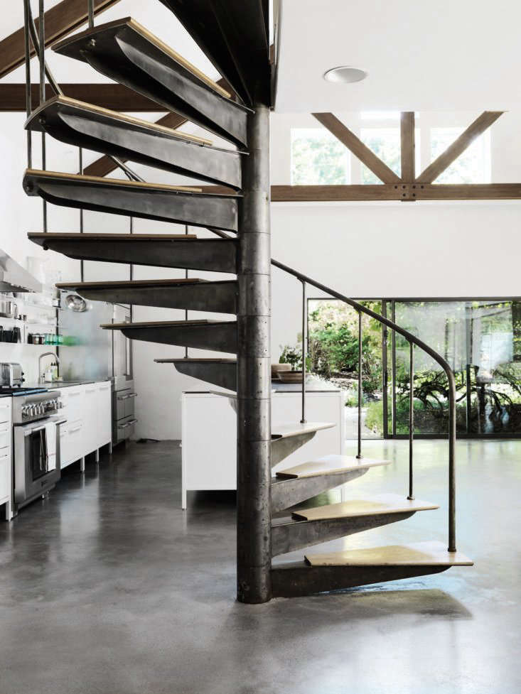 The kitchen is in a new extension of the house that leads to an outdoor patio. The steel spiral stair, polished concrete floor, and barn-style joists stand out against bright white walls.