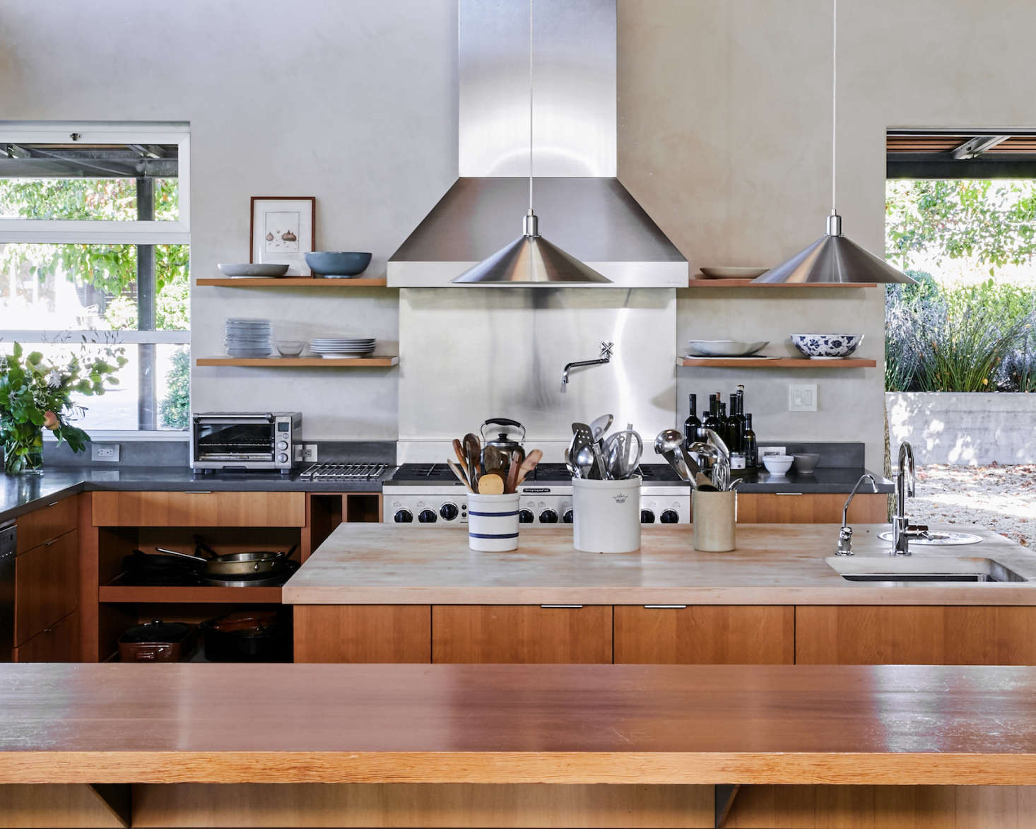 The large central island has a butcher block countertop, integrated sink and compost pail, and both open and closed storage on its sides. The breakfast bar in the foreground is made of solid Douglas fir. The doorway to the right leads to an outdoor pea gravel patio with dining table and wood-fired pizza oven, where wine tastings are held in good weather.