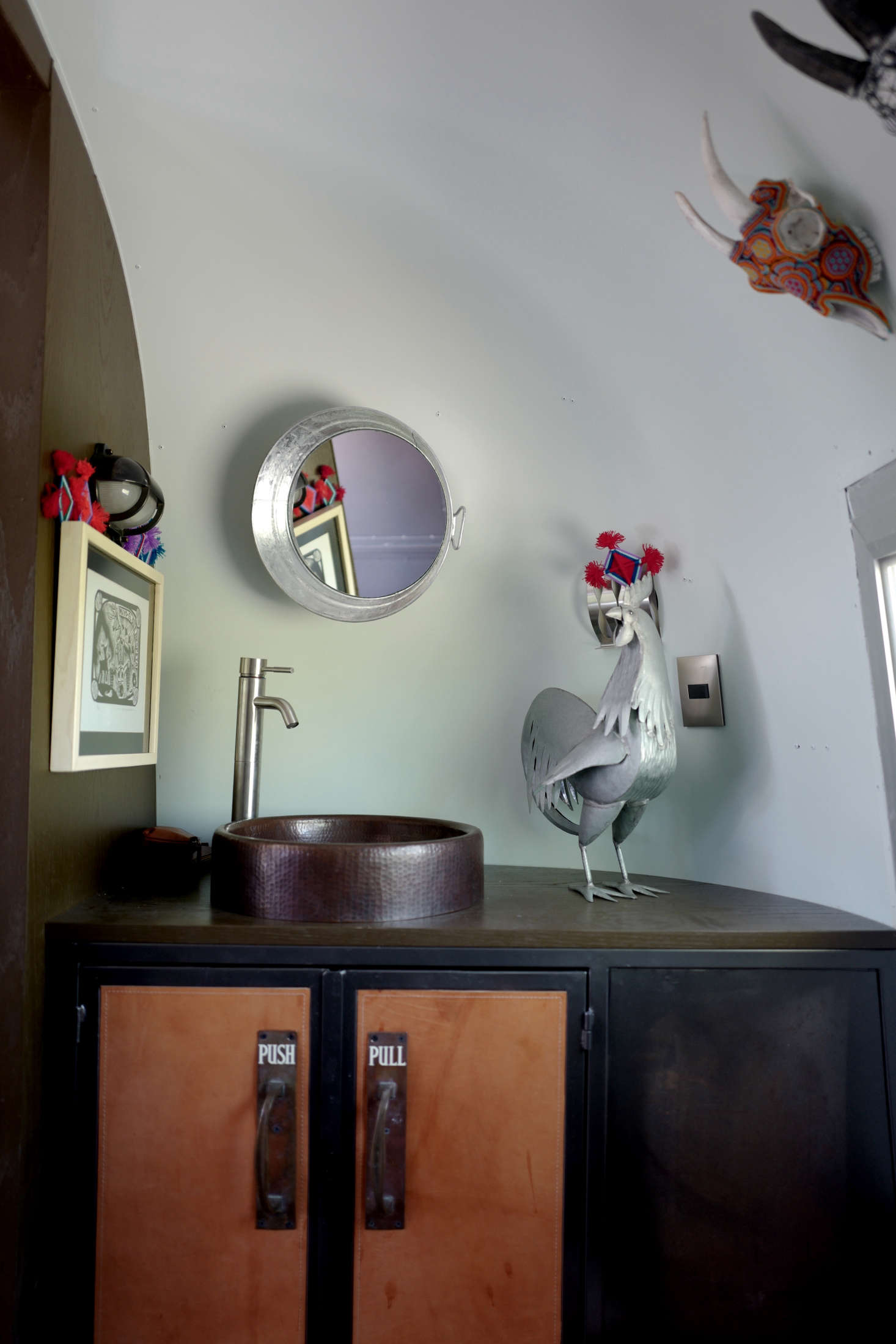 The compact bathroom has a leather-detailed iron vanity with a hammered copper sink. The couple found the vintage cabinet pulls in Arundel, England. The galvanized tin rooster is the work of one of the Casamidy artisans. Also made of galvanized tin: the washtub mirror.