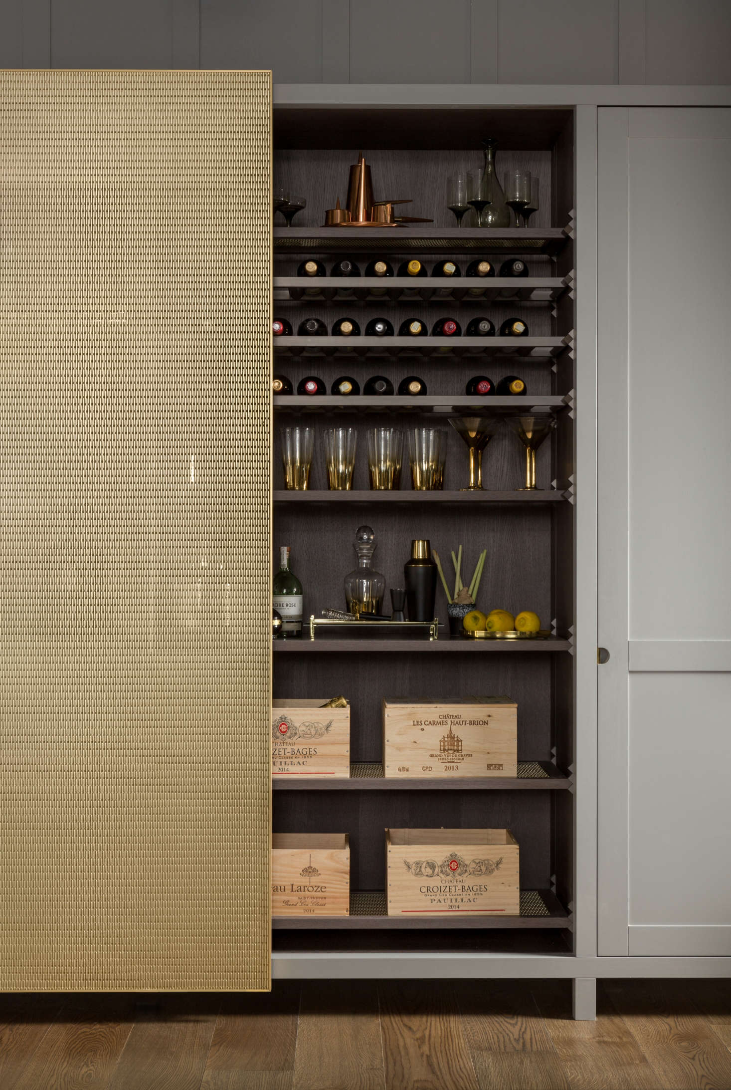 Behind the sliding brass mesh door is a wine section detailed with Lanserring’s sawtooth shelf supports. It has bottle racks, leather-topped glassware shelves, and brass-mesh-inset shelves for ventilation.