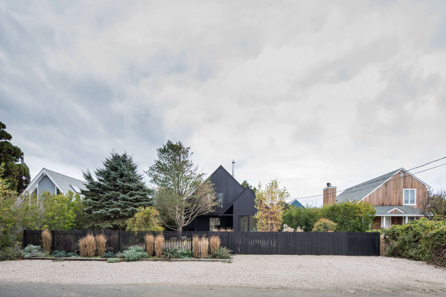 The beach house is clad in cedarwood, stained with eight coats of Black Cabot stain. Two four-foot-tall fences define the property from the street. The fence on the right is almost solid, to hide the streetside gravel parking court from ground-floor occupants. The slatted fence on the left lets light in and “allows the native grasses and dunes flow through.”