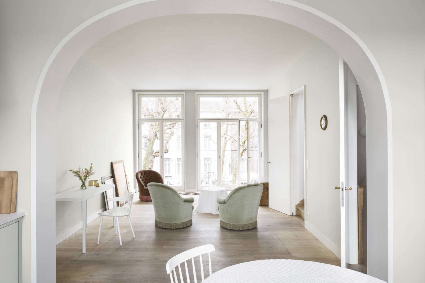 Looking back toward the front of the townhouse, with the tops of the plane trees showing through the windows and the inset staircase visible at right. These photographs were taken when the homeowner was moving back in; the velvet chairs in sage and rust are the architects’ own, and the small round table, draped with a cloth, is a placeholder.