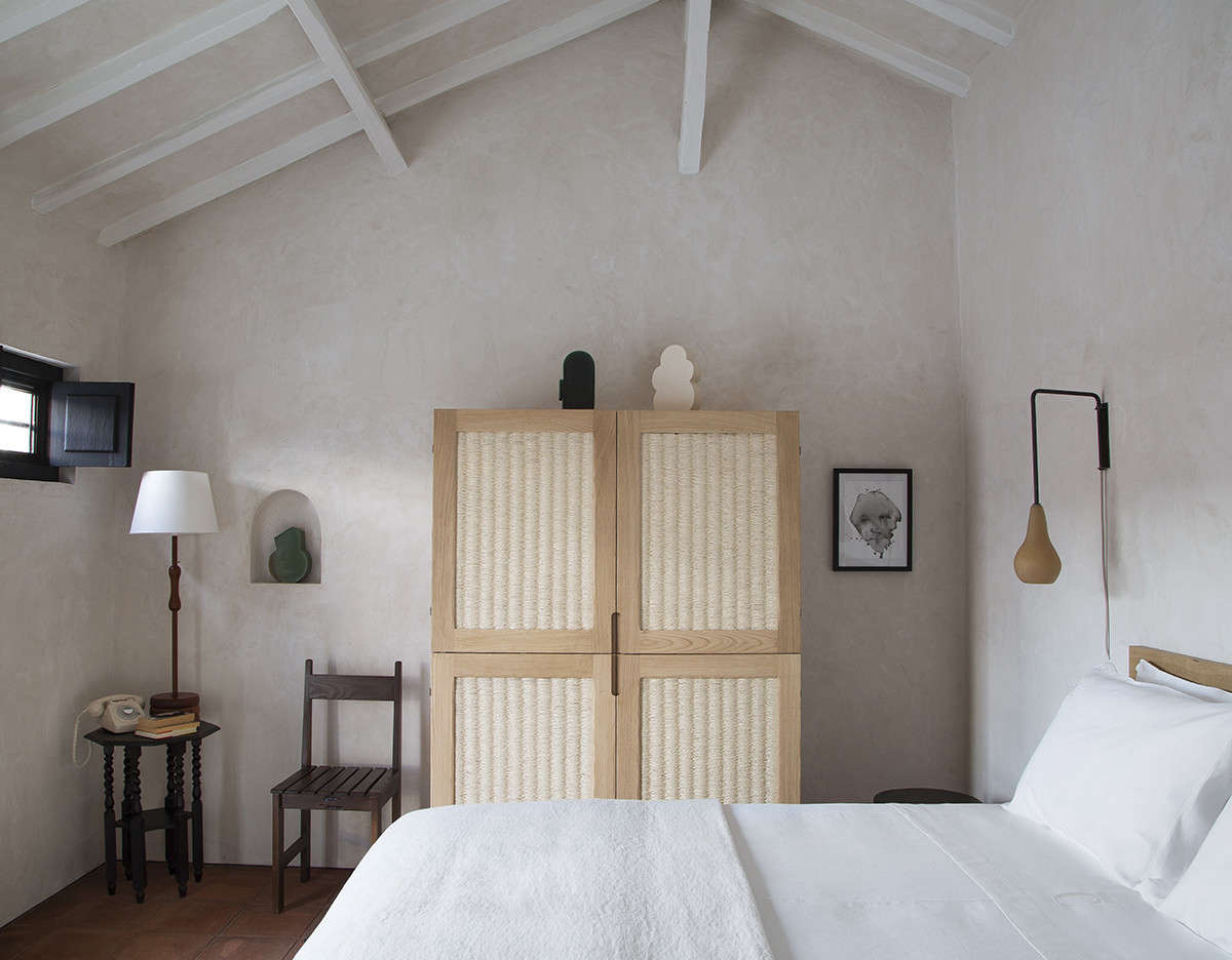 The eight guest bedrooms are coated in plaster, so that even empty walls feel textural. Note the sense of balance here, too, with a pear-shaped sconce weighted against the small black-painted window on the other side of the room, and a chair and tall lamp on either side of a recessed niche.