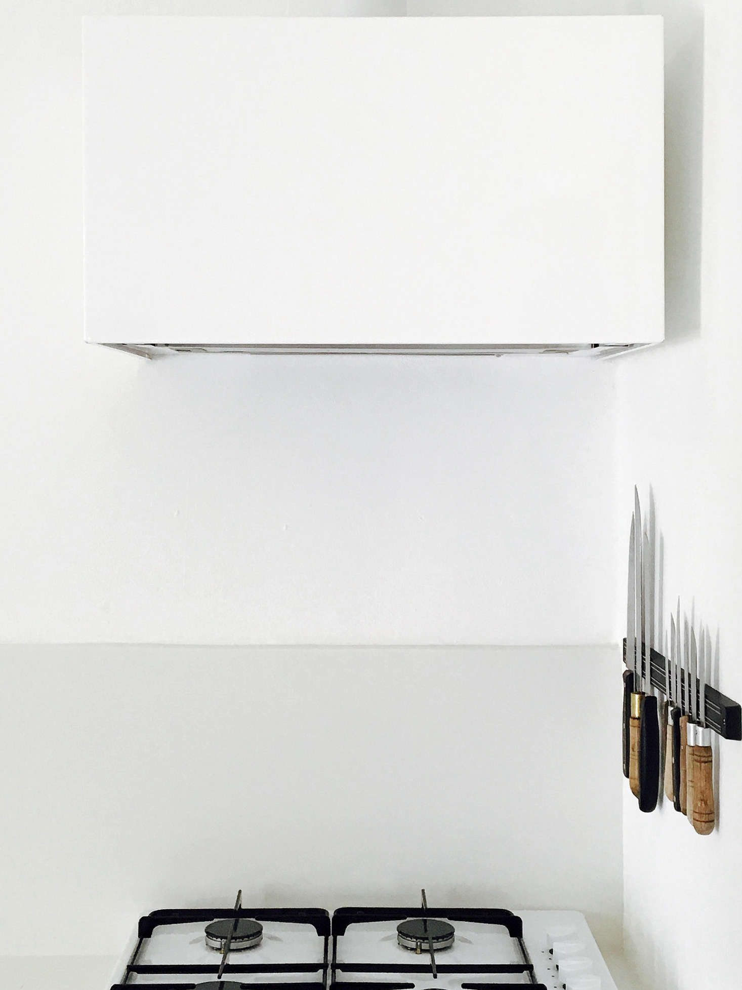 In an otherwise austere kitchen, necessary storage doubles as design, as in the knife rack beside the cooktop. The sculptural vent hood cover and the base for the range are both made of thin steel, painted with primer and matte white paint, and made by a local metalworker.