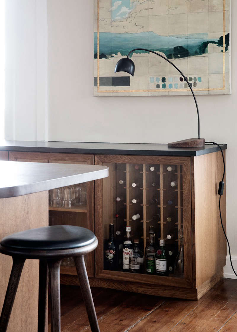A combination bar and storage cabinet stands at the other end of the kitchen, matched by a pair of glass-fronted storage cabinets near the sink. The leather-topped High Stools are by Space Copenhagen.
