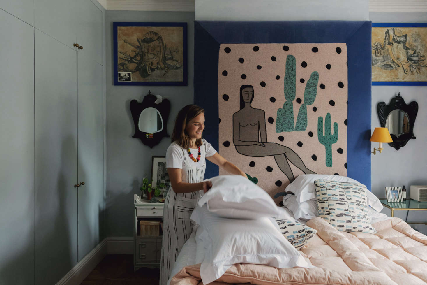Heuman tidying up her bedroom. “Good storage is essential. I love organizing my home—I call it a ‘streamlining sesh’—and it’s so satisfying once everything has been put away. It lifts the whole environment,” she shares. (To learn more about how she keeps organized, read her tips in the NY Times.) A charming woven throw blanket by BFGF that she had framed in blue velvet makes up the headboard.